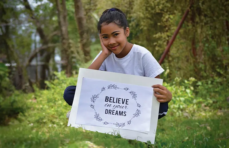 Young girl sitting in forest, holding a sign that says 'Believe in your dreams'